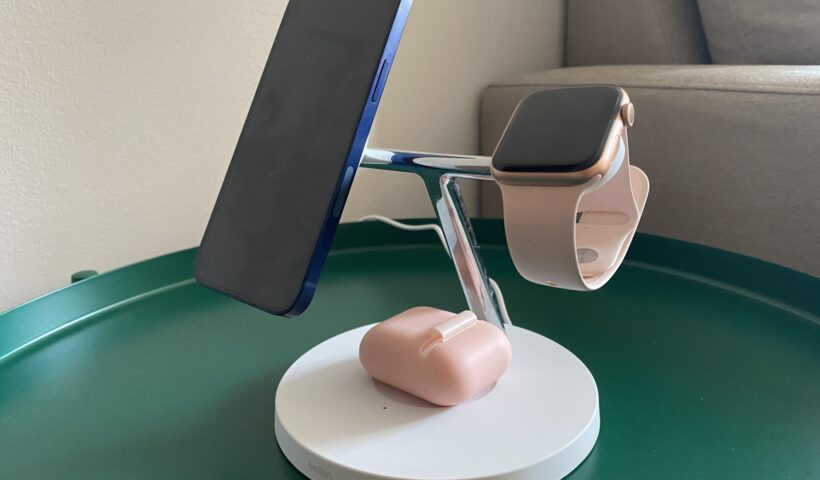 A white 3-in-1 wireless charging stand with an aluminum T-bar design, holding a blue iPhone 12, pink Apple Watch, and AirPods in a pink case.