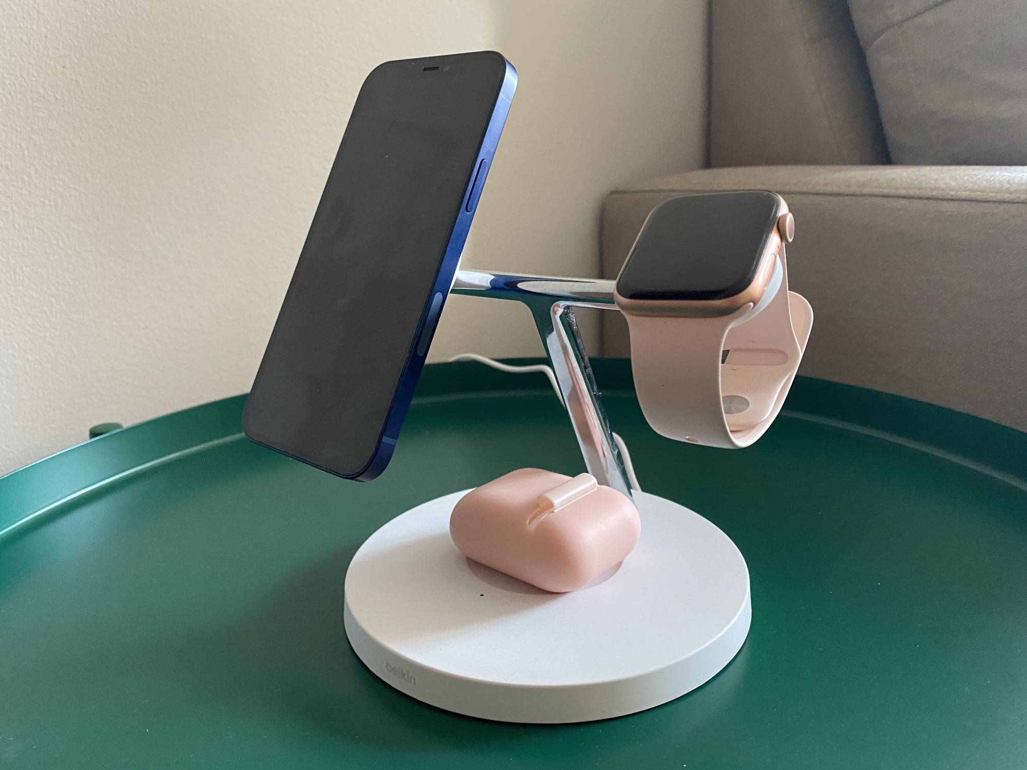 A white 3-in-1 wireless charging stand with an aluminum T-bar design, holding a blue iPhone 12, pink Apple Watch, and AirPods in a pink case.