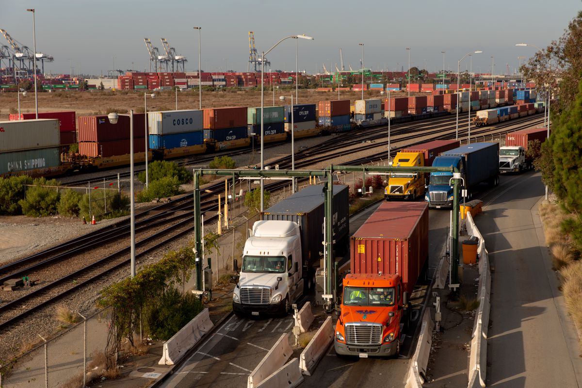 Large trucks are transporting shipping containers on a road. In the distance, there are more shipping containers.