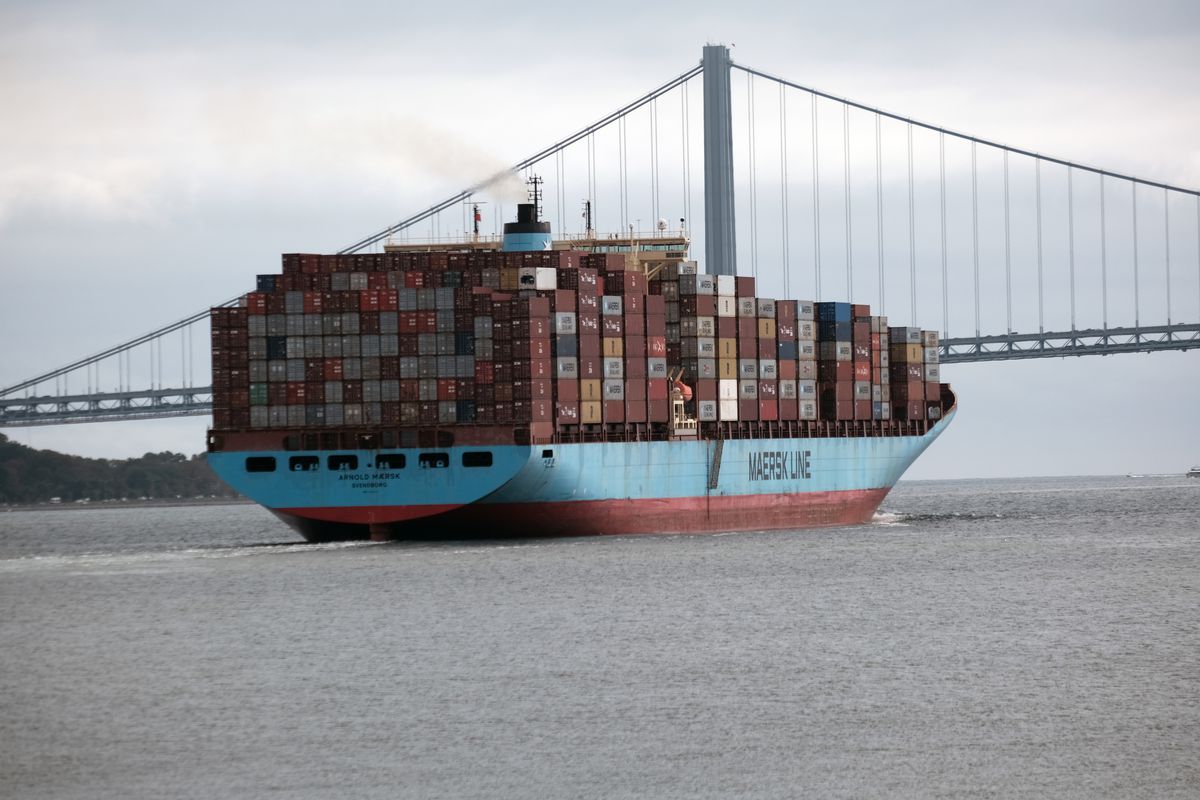 A shipping container ship, full of shipping containers, on the water in front of a bridge.