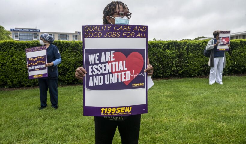 Health care workers outside a care facility hold signs that read, “Quality care and good jobs for all. We are essential and united. 1199 SEIU.”