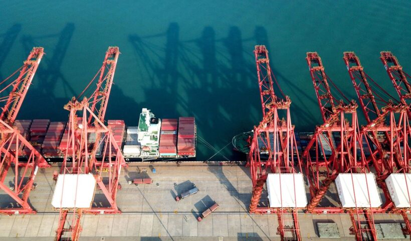 Large red cranes are seen next to trucks and a ship, which is full of shipping containers.