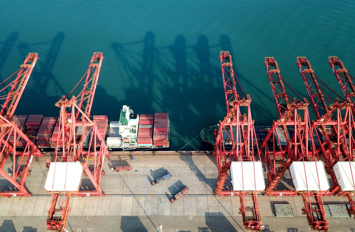 Large red cranes are seen next to trucks and a ship, which is full of shipping containers.