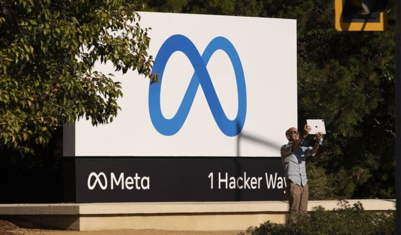 A person stands in front of the Meta company sign at 1 Hacker Way taking a selfie.