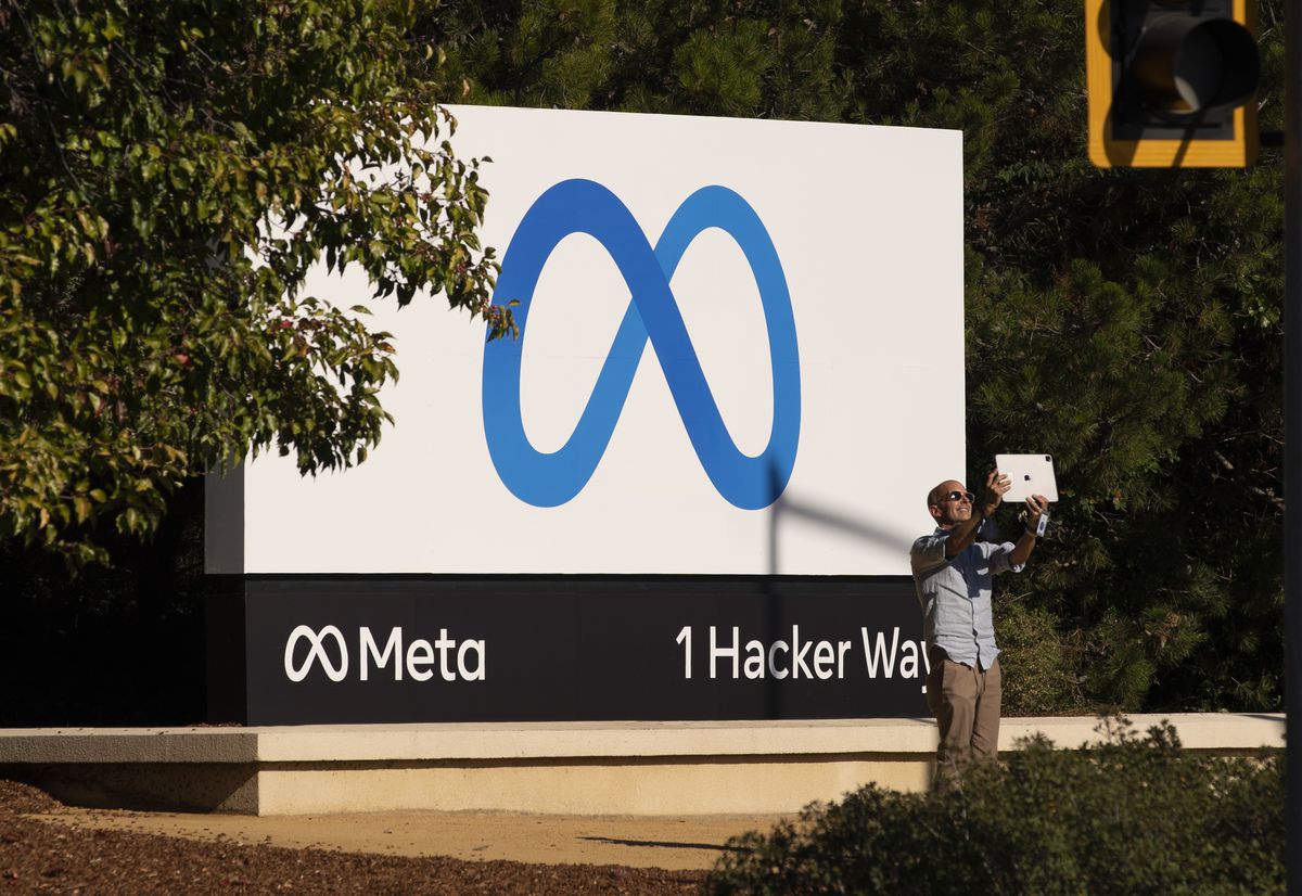 A person stands in front of the Meta company sign at 1 Hacker Way taking a selfie.