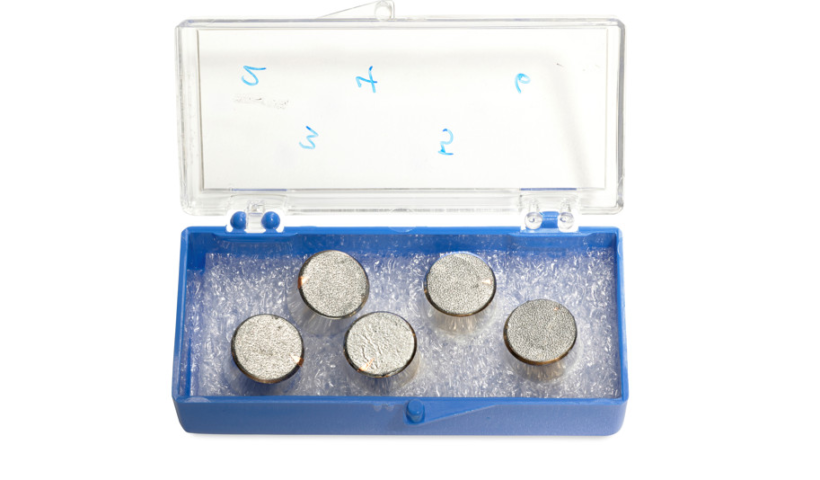 An open sample box containing five metal discs, each with moon dust stuck to it.