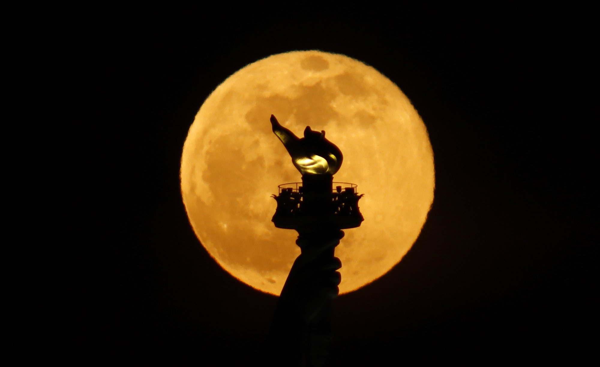 The Statue of Liberty's flame silhouetted against a yellow-tinged full moon.
