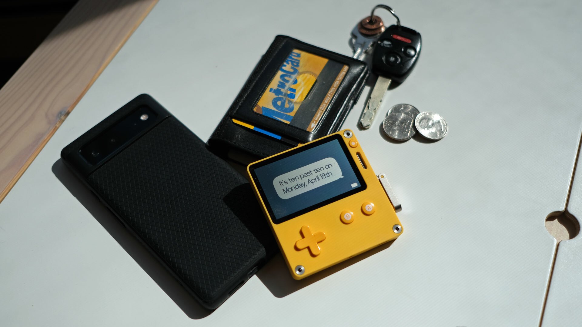 A Playdate gaming device seen from above, surrounded by a phone, wallet, keys, and coins.