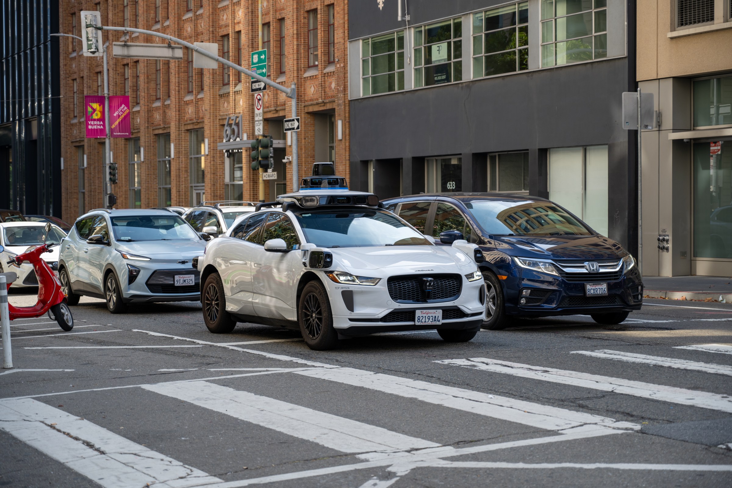 A white self-driving car equipped with sensors and cameras on the roof waits at an intersection in a city, surrounded by regular vehicles, including a silver hatchback and a dark blue sedan. A red scooter is parked on the side of the street, and brick and glass buildings line the background.